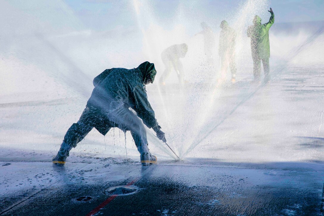 A sailor adjusts a sprinkler as water sprays on the deck of a ship; others stand in the background.