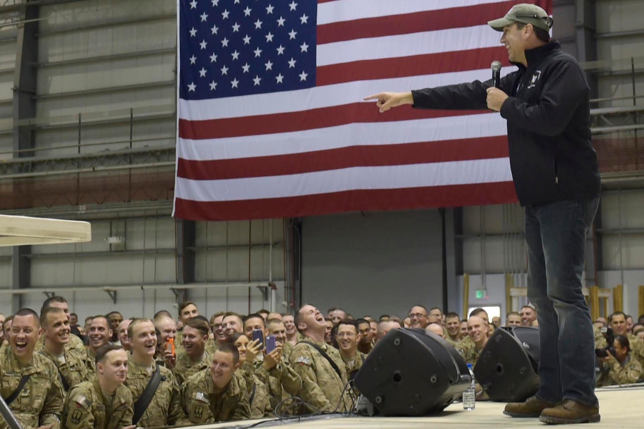 A man performs on stage before a group of service members; a giant U.S. flag is on the wall to the right.