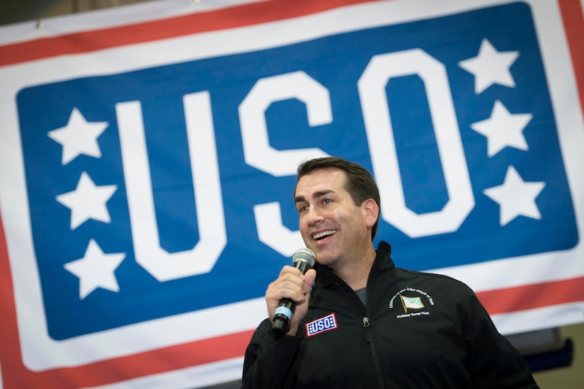 A smiling man holds a microphone; behind him, a banner spells out USO.