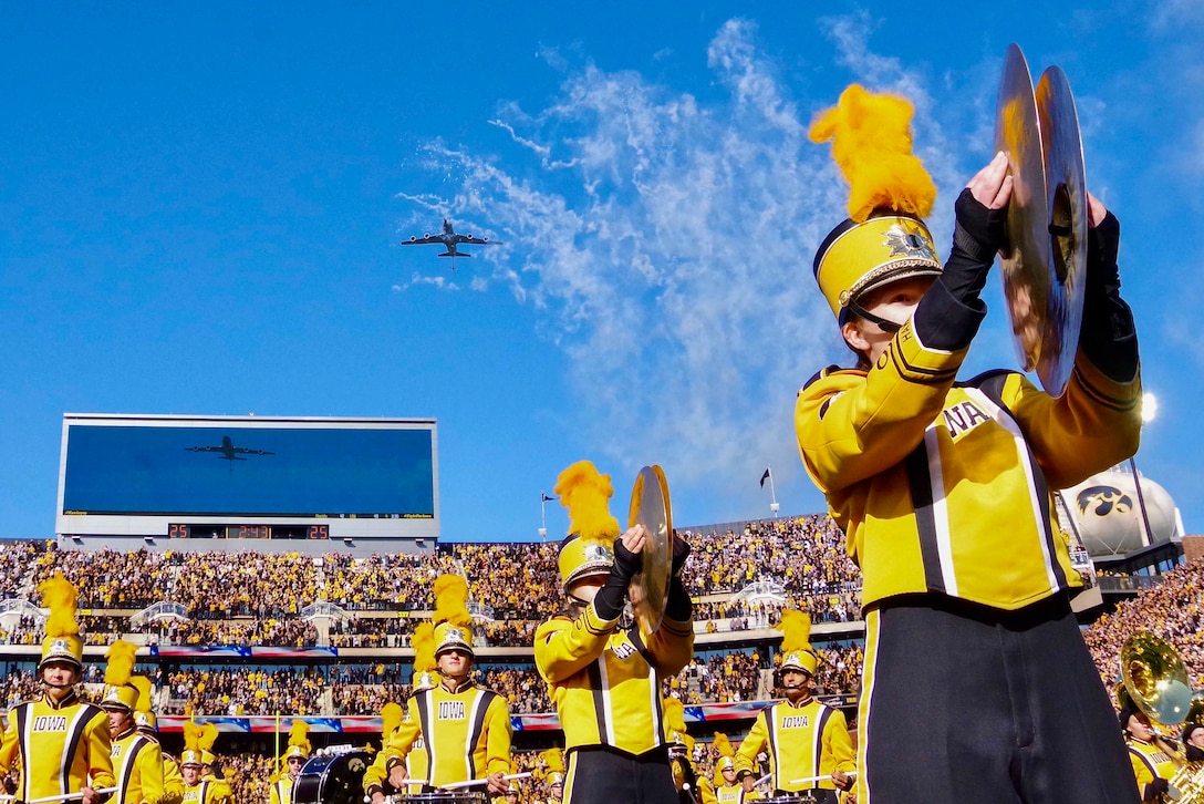 A military aircraft flies over a stadium as a band performs.