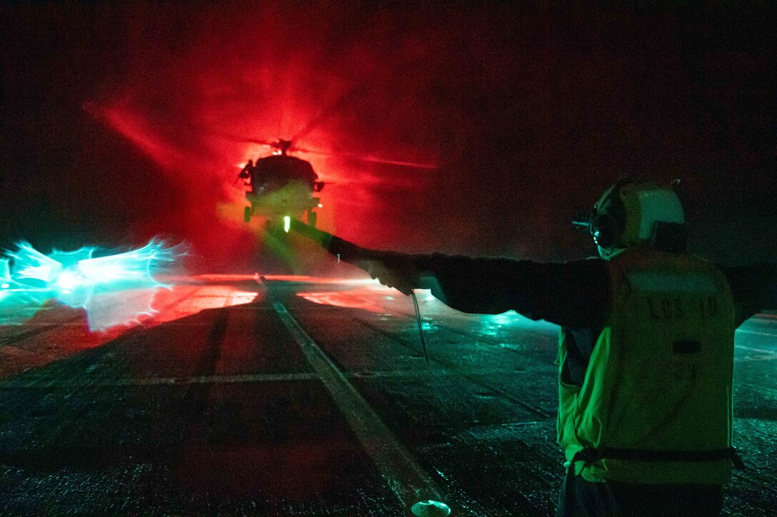 A sailor signals a helicopter on the flight deck of a military ship at night.