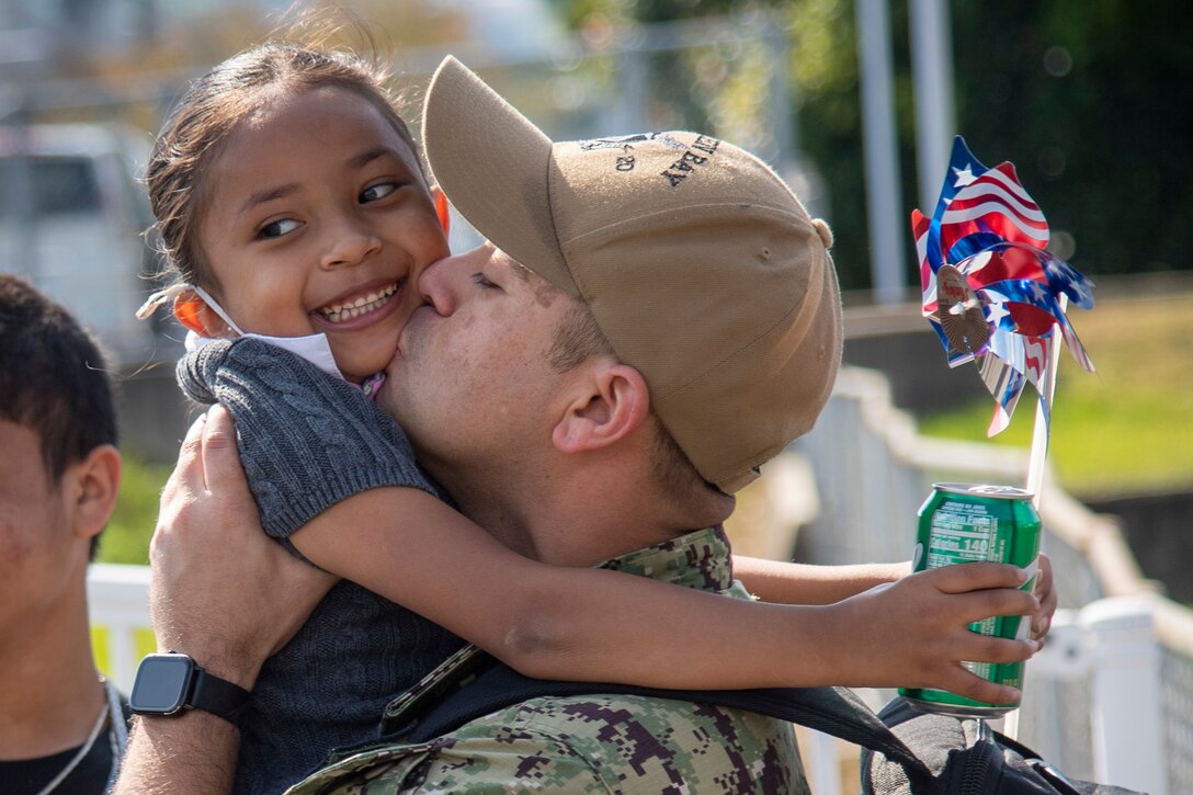 A sailor holds his daughter in his arms and kisses her on the chin.