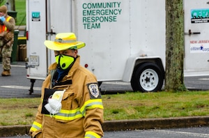 A male first responder in a yellow hat, face  mask and jacket stands outside in front of a white truck with the words "First Responder Team" printed on the side.