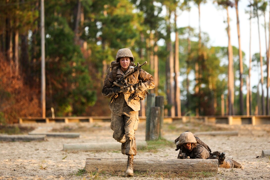 A Marine Corps recruit runs on an obstacle course while a fellow Marine lays on the ground and aims a weapon.