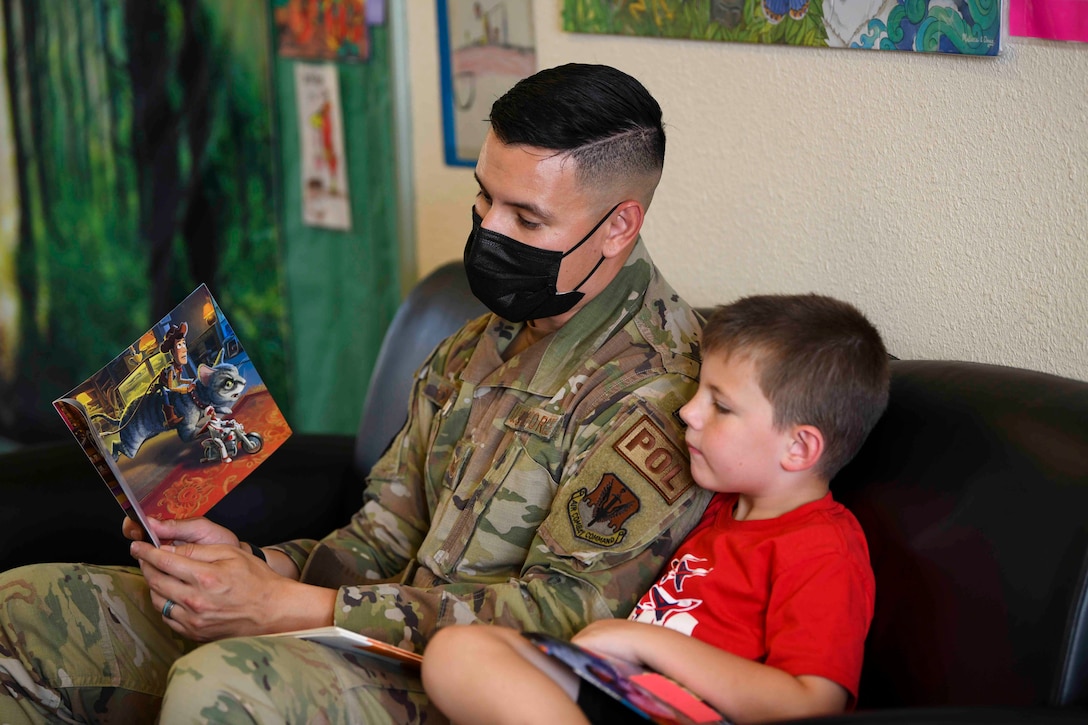 An airman sits on a couch reading a book to a child.