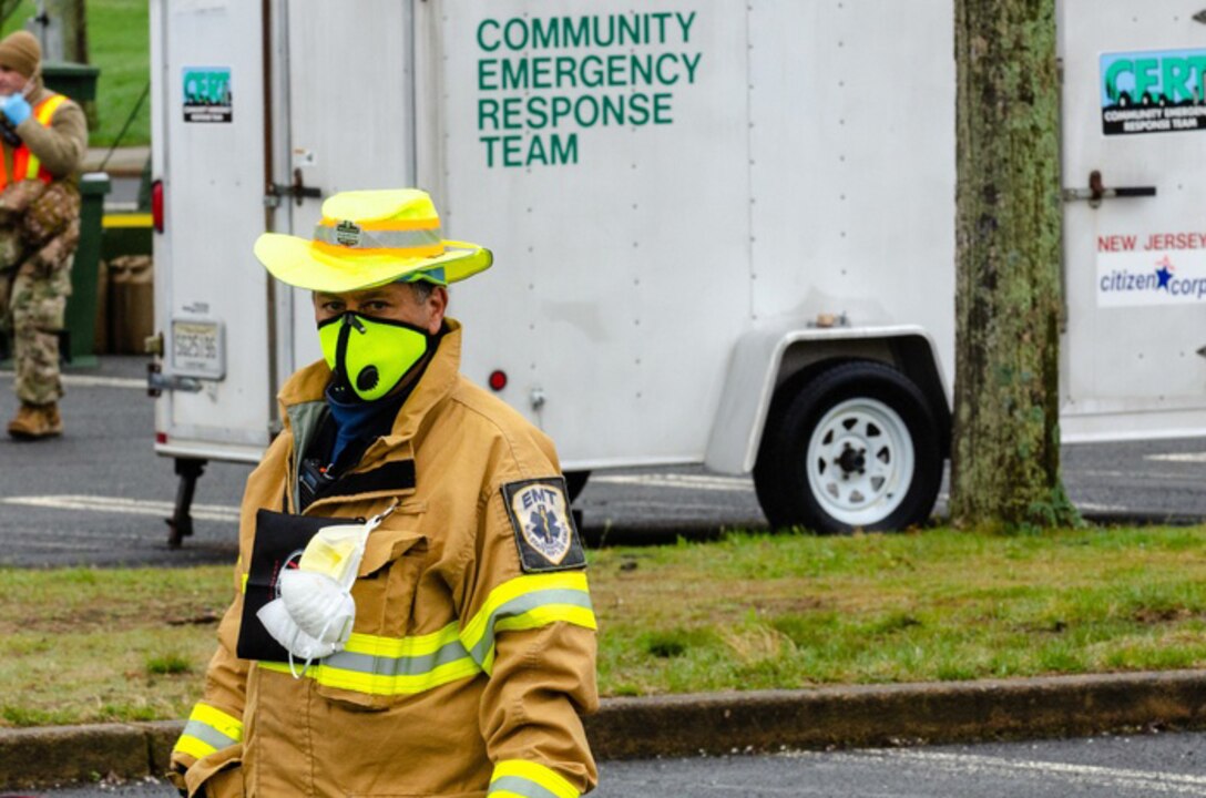 A first responder in a yellow hat, face  mask and jacket stands in front of a white truck with the words "Community Emergency Response Team" printed on the side.