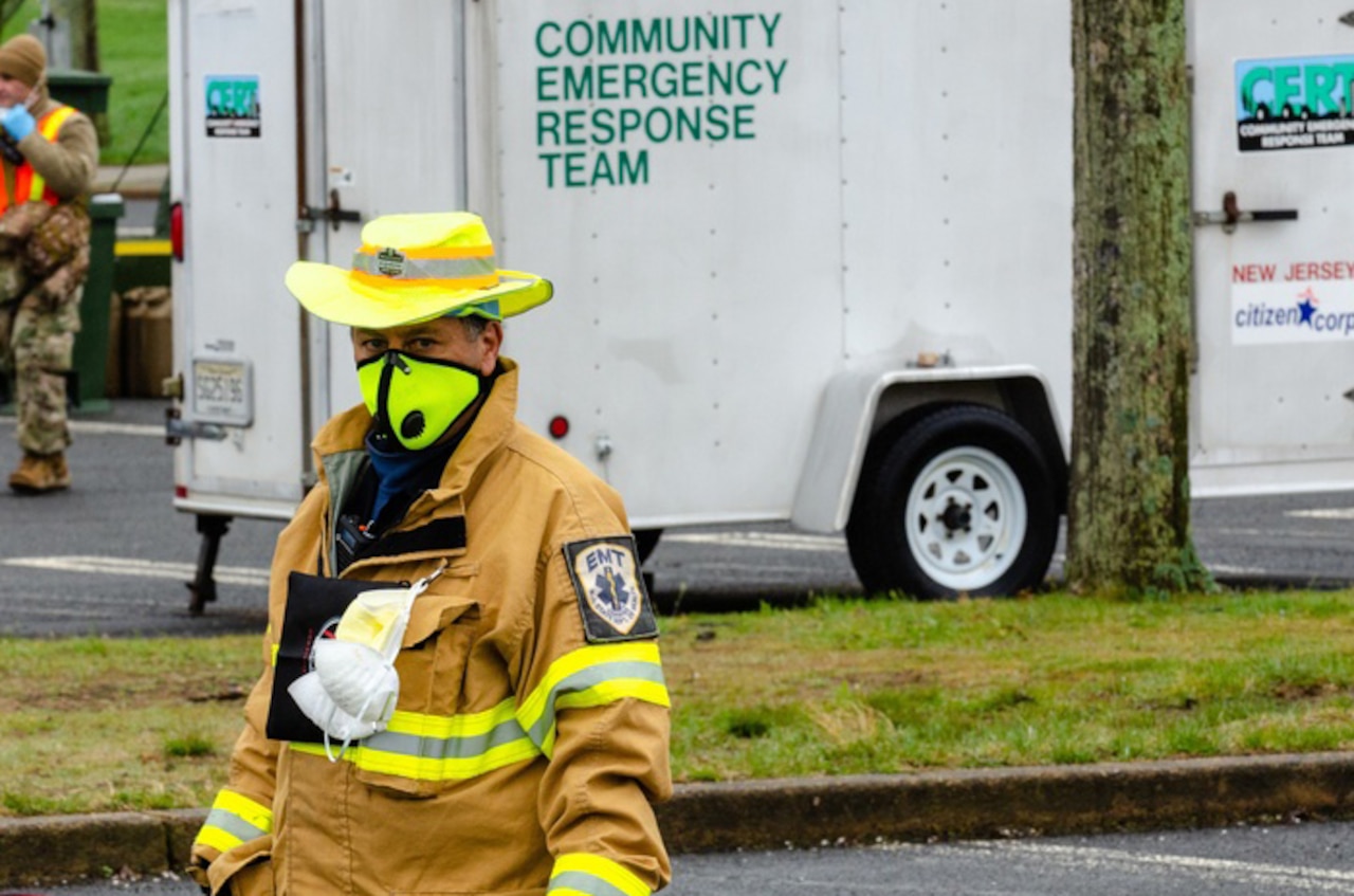 A first responder in a yellow hat, face  mask and jacket stands in front of a white truck with the words "Community Emergency Response Team" printed on the side.