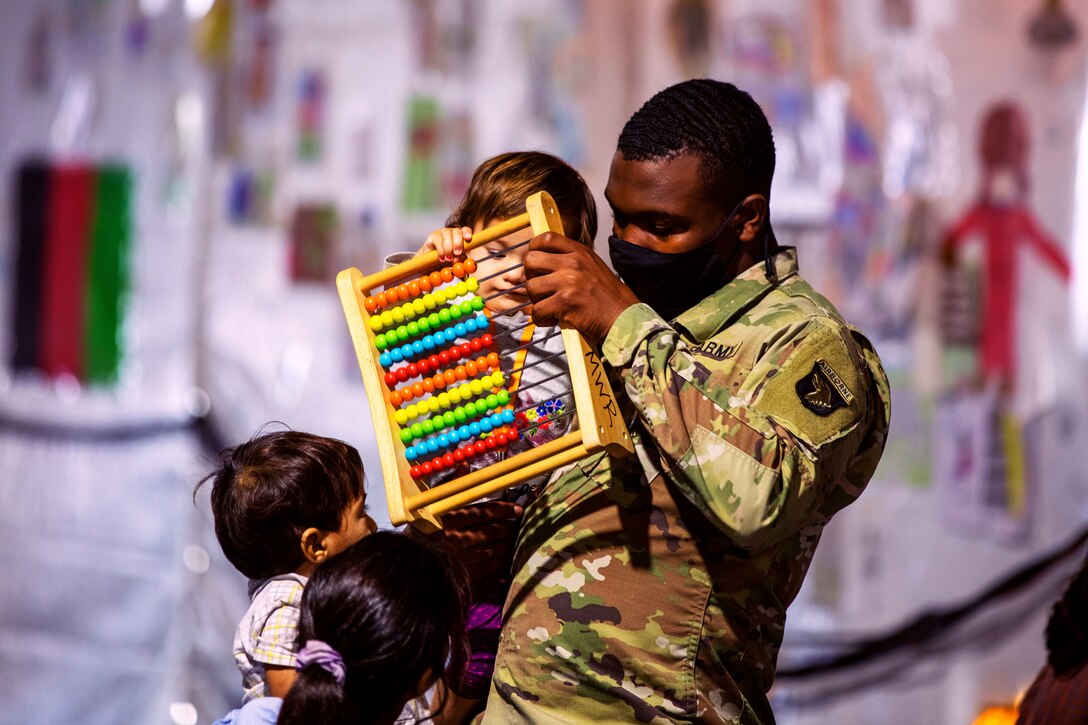 A soldier wearing a face mask holds and plays with one Afghan child while two other Afghan children stand in front of him.