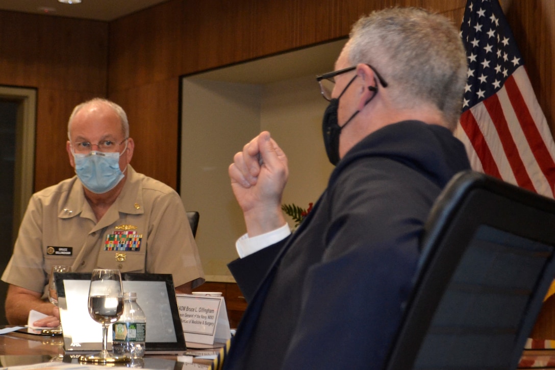 A man in Navy uniform looks intently across a table at another man in a suit as he speaks.