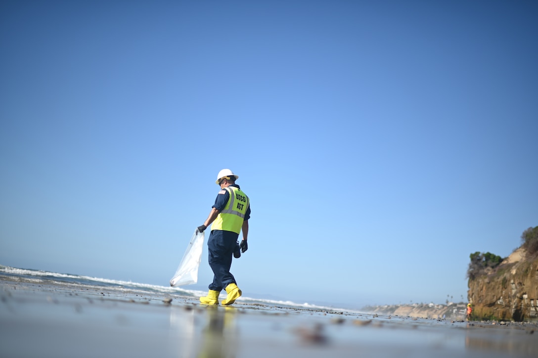 A man carrying a plastic bag looks for oil tar balls on a beach.