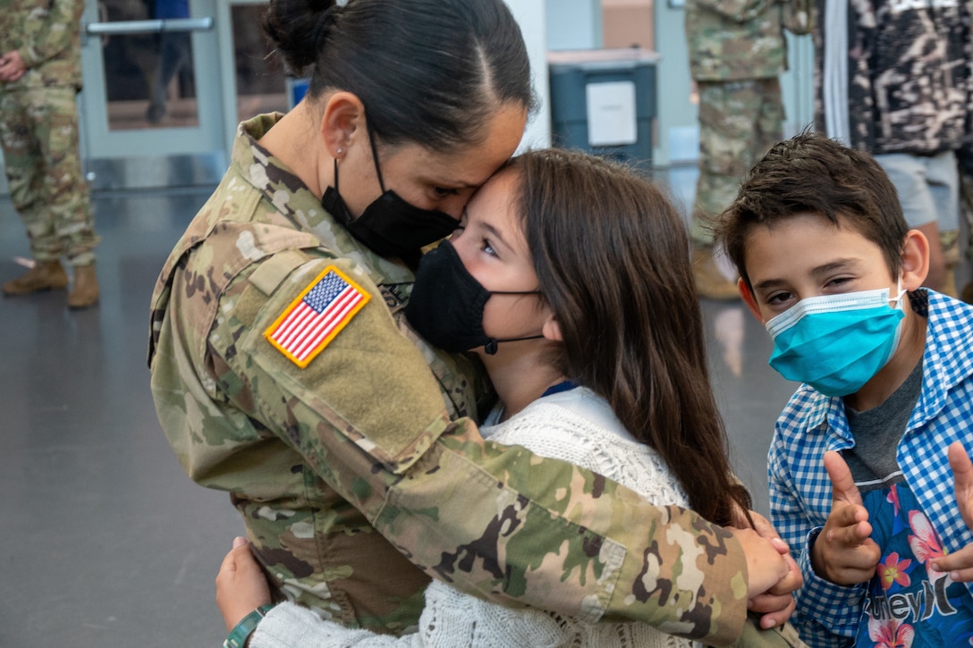 A soldier hugs her daughter while her son gestures and smiles at the camera.