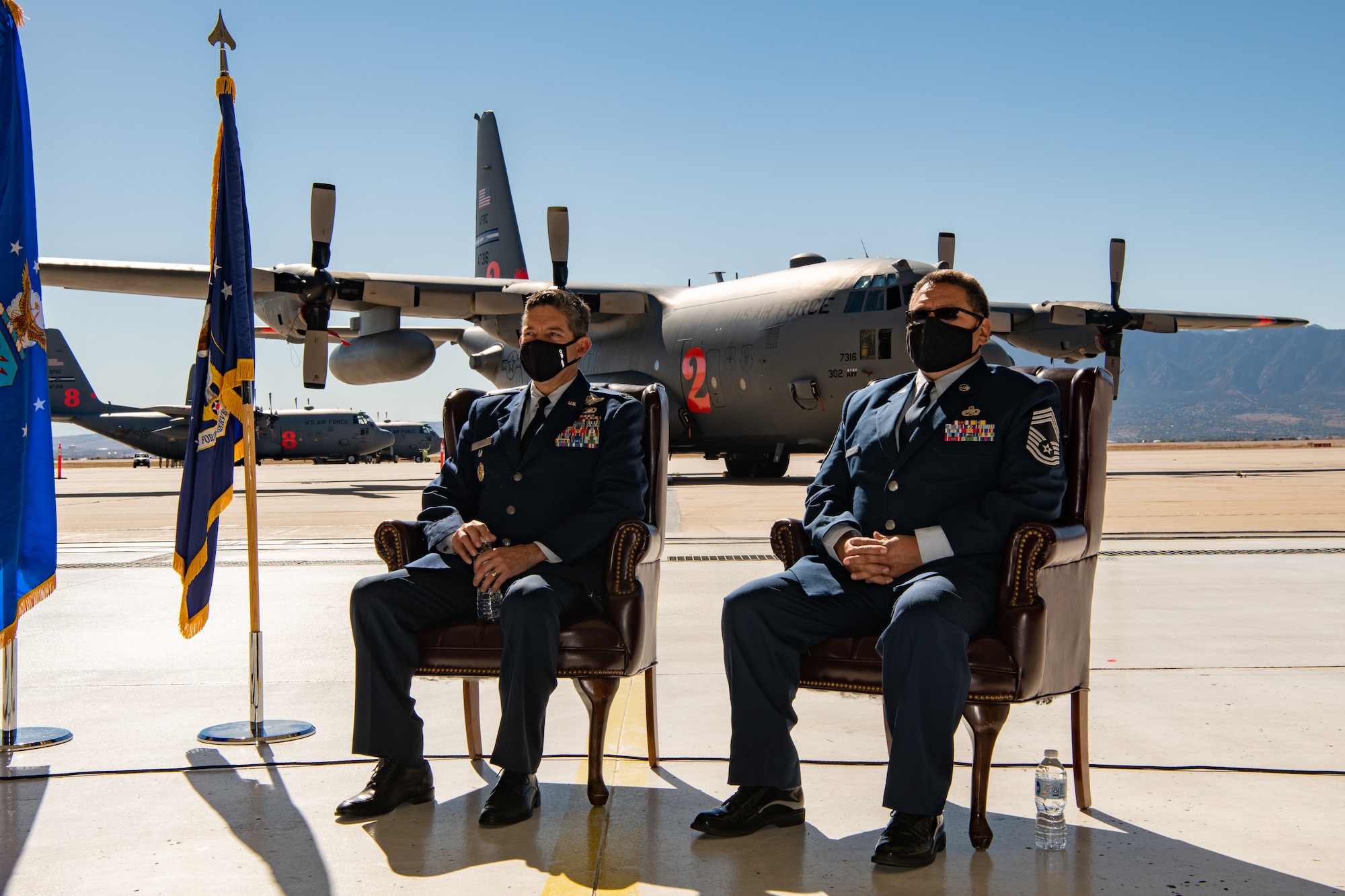 Two men in service dress sit in chairs with a C-130 on a parking ramp behind them.