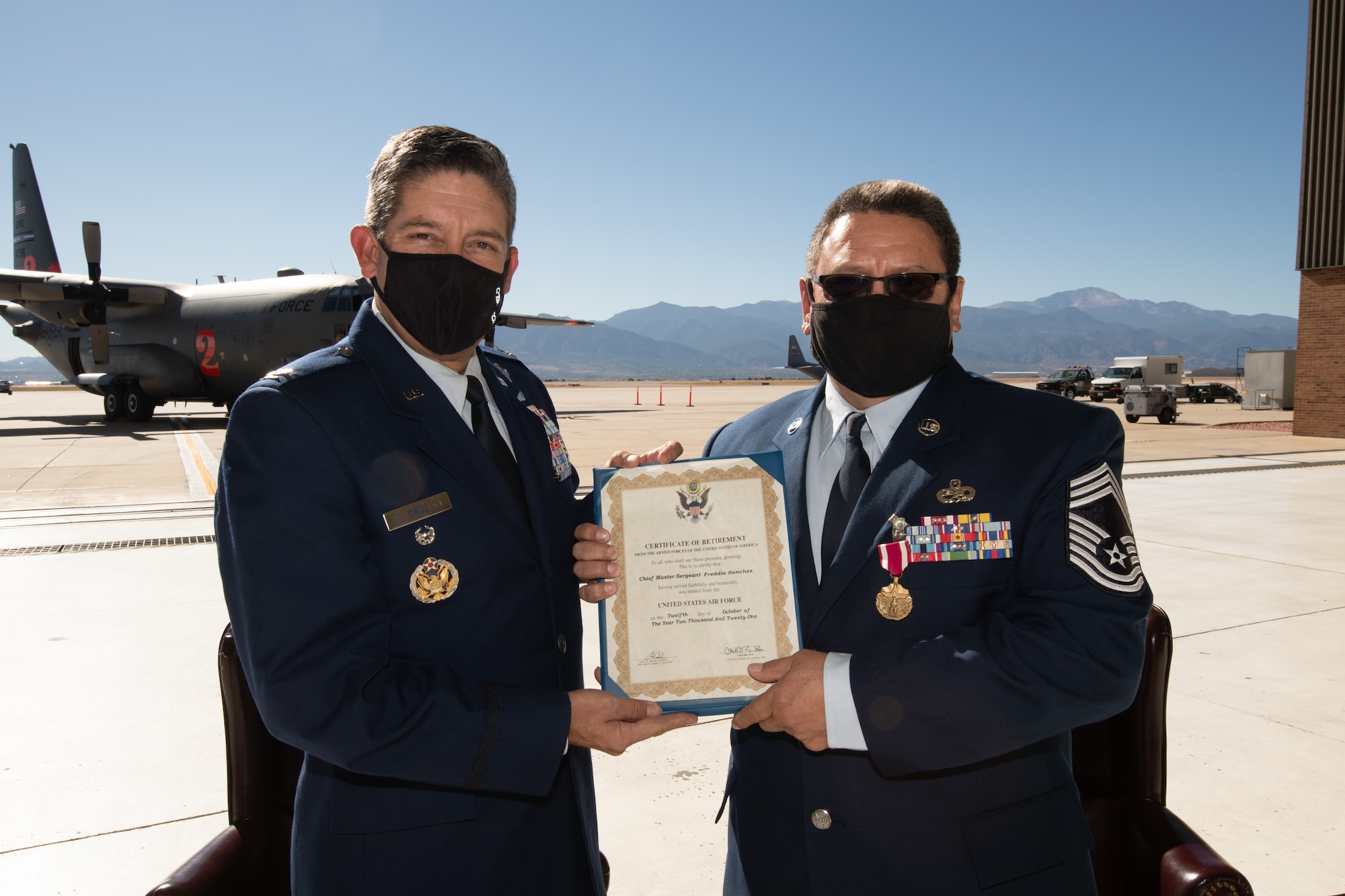 Two men in Air Force service dress stand in a maintenance hangar with a retirement certificate between them. In the background is a C-130 on a parking ramp and a mountain range in the distance.