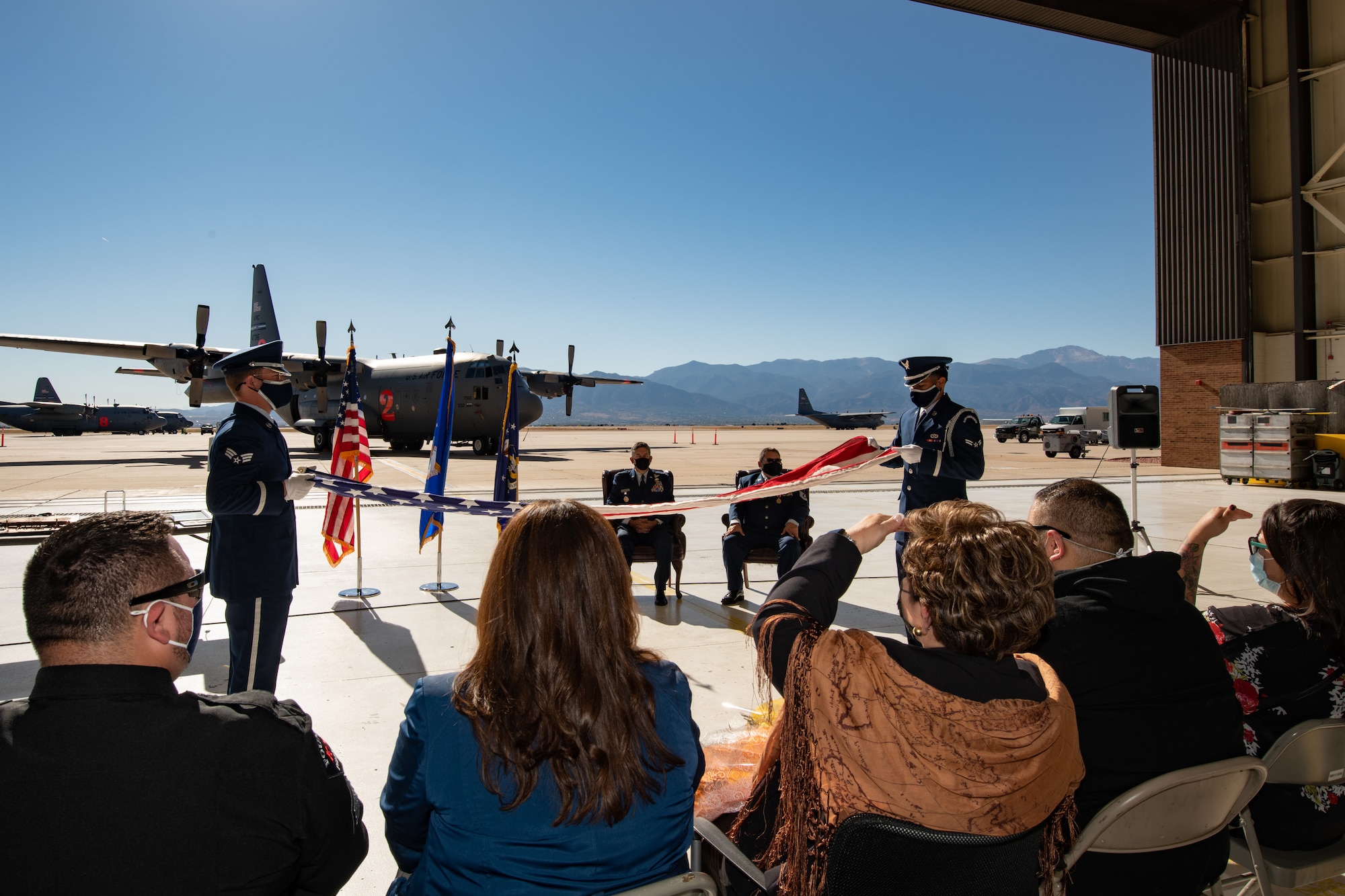 Two Airmen in service dress unfold an American flag in front of two men in Air Force service dress with two C-130s on a parking ramp behind them and a mountain range in the background.