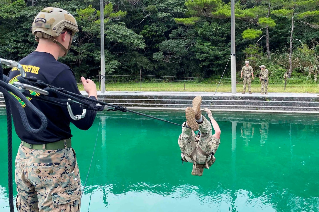 A soldier crosses a rope bridge over a pool as others watch.