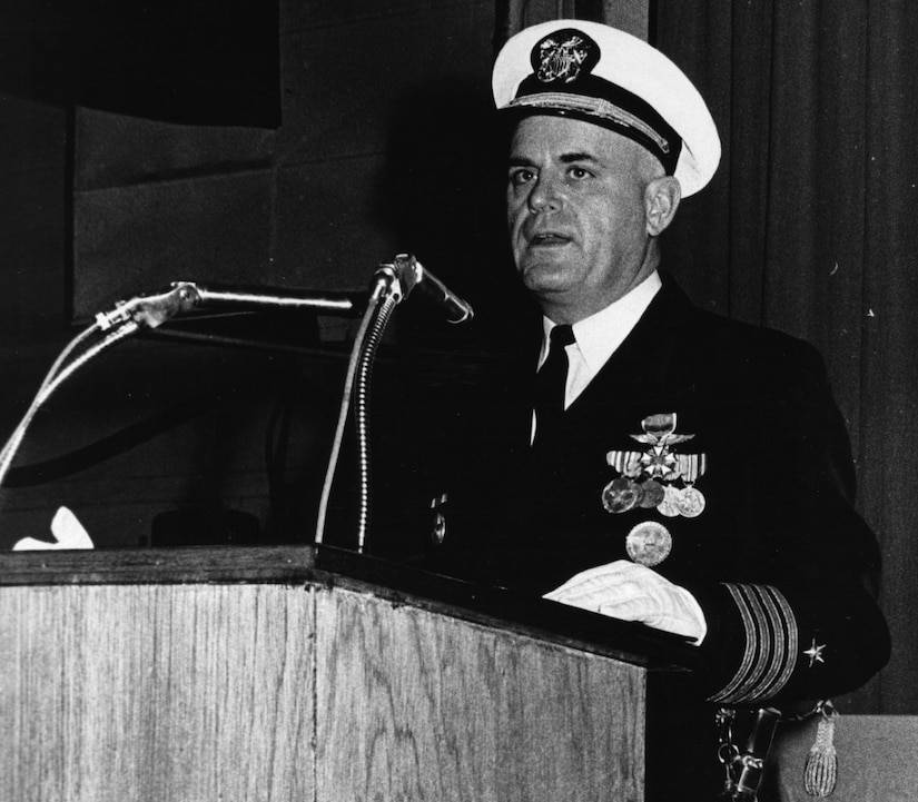 A man in a dress uniform and cap stands at a podium.