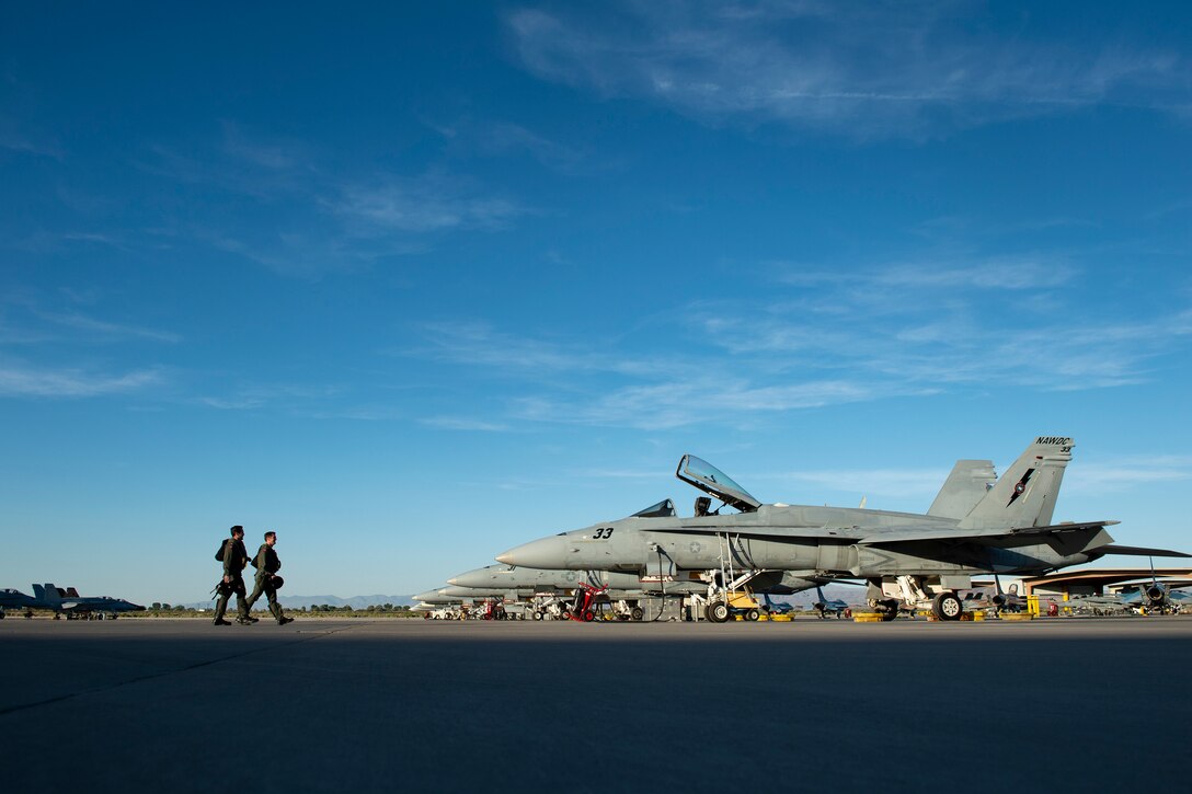 Two men walk along an airstrip as large jets sit in front of them under a blue sky.