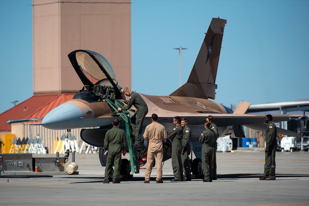 A pilot walks down a ladder attached to the cockpit of a fighter jet. Several people stand below him on the tarmac.
