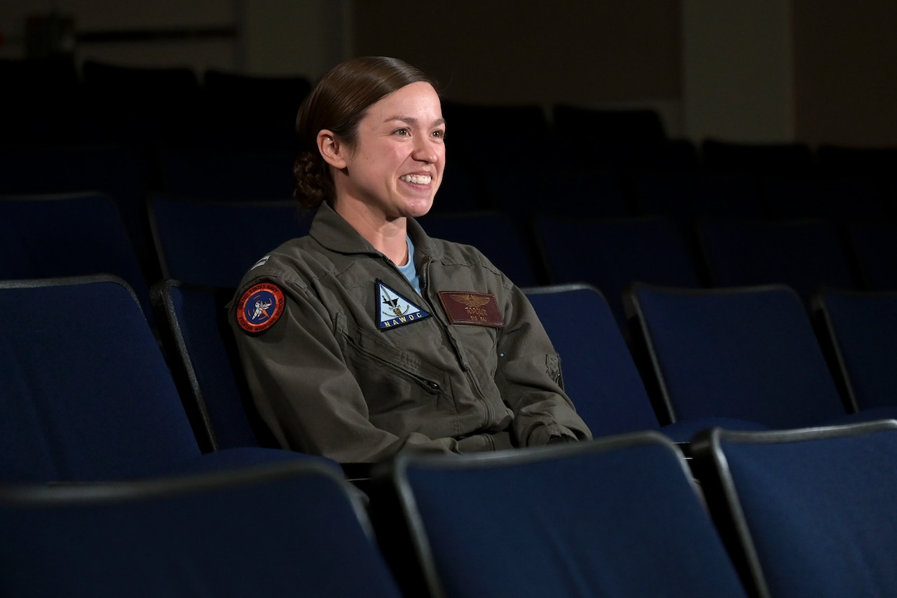 A woman in a flight suit smiles while sitting in an auditorium chair.
