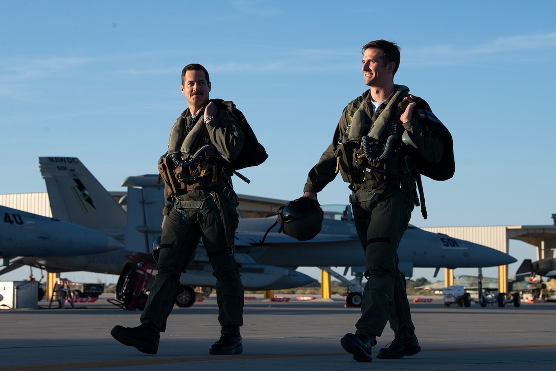Two men in flight gear walk on tarmac with jets in the background.