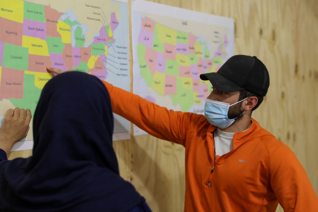 A man wearing a face mask and baseball cap points to Kansas on a map of the U.S. while speaking to an Afghan woman.