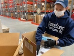 Guy in blue sweatshirt, blue ball cap and white face mask arranges items in a box.