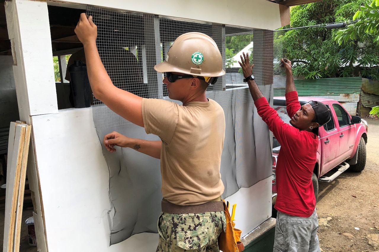 A service member and civilian install a window screen on a building under construction.