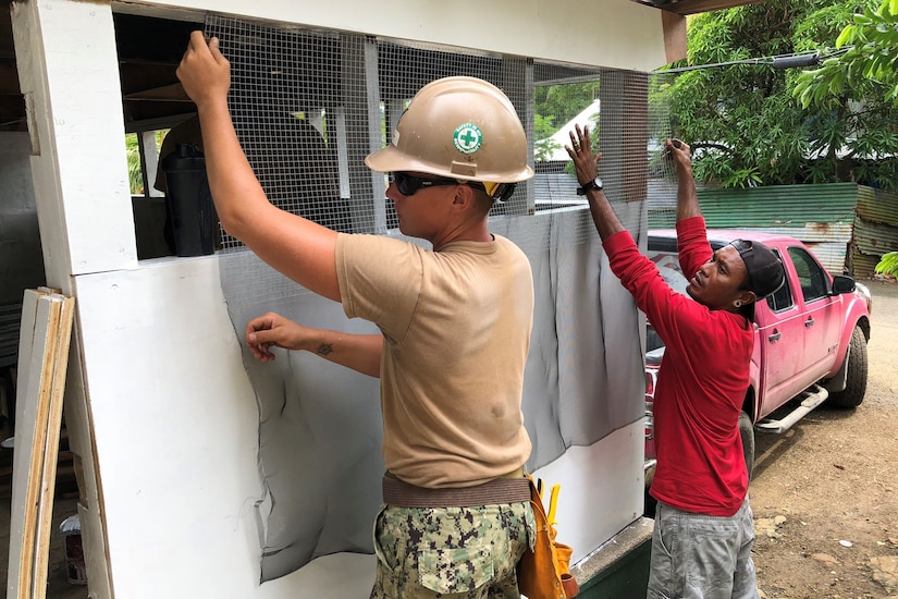 A service member and civilian install a window screen on a building under construction.