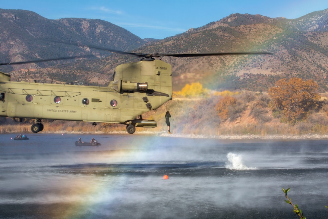 A soldier jumps out of a helicopter hovering over a body of water as seen through a rainbow.