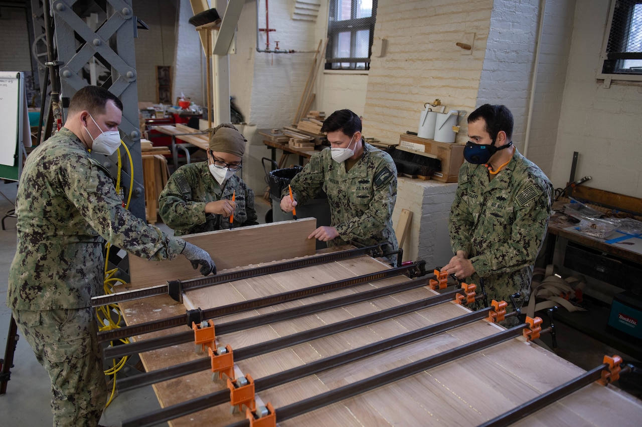 Four sailors work on unfinished wood planks on a table.