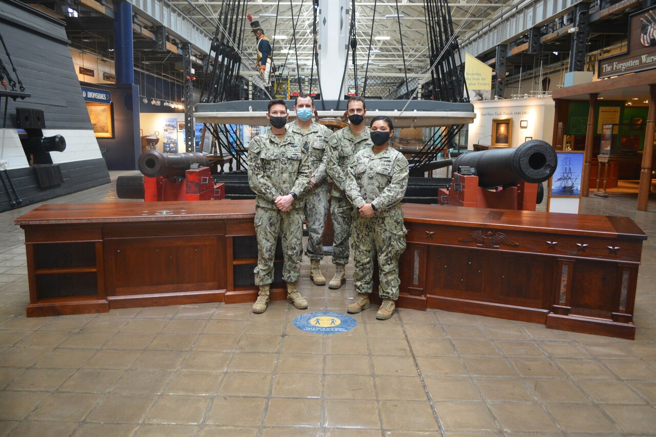 Four sailors stand between two wood desks in a museum.