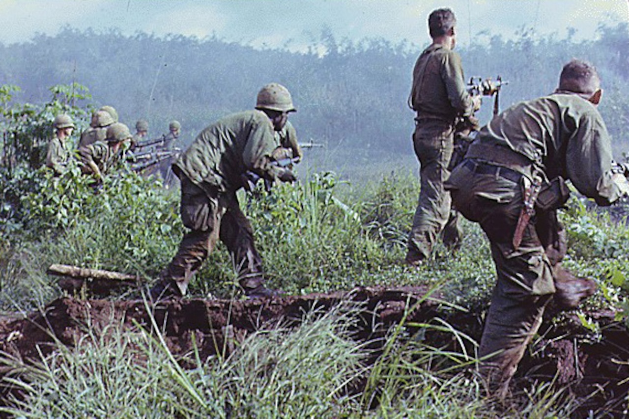 Three soldiers with rifles duck in jungle brush while looking toward trees. Other men are doing the same in the background.
