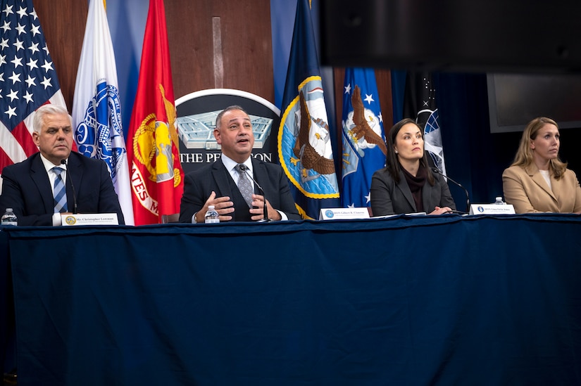 Two men and two women sit at a table during a committee meeting. Multiple flags are behind them.