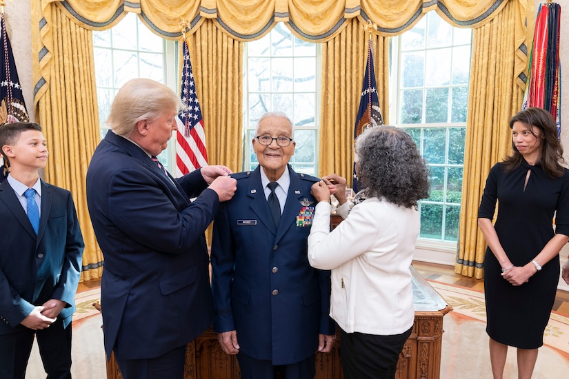 A man in military uniform is flanked by two civilians. Their hands are on his shoulders.