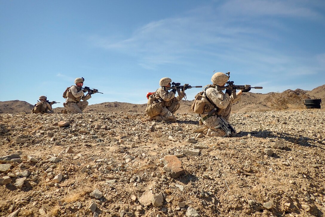 Four Marines kneel in a desert and aim weapons.