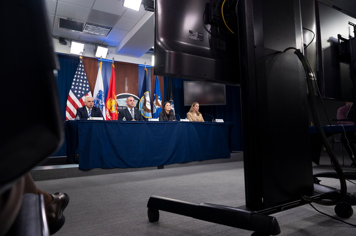 Two men and two women sit at a table with the U.S. flag and four service flags behind them.