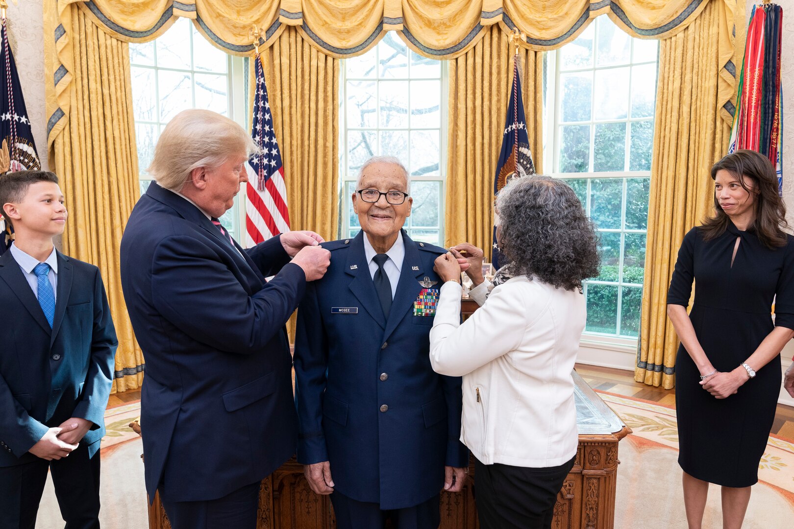 A man in military uniform is flanked by two civilians. Their hands are on his shoulders.