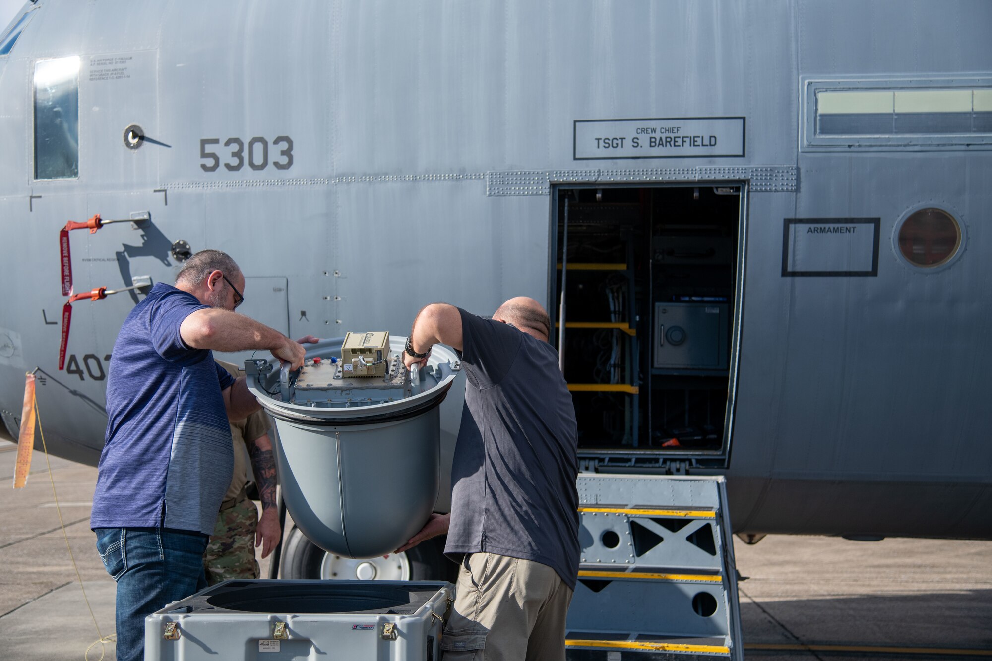 Two men lift a satellite radome from a plastic bin. Aircraft in background.