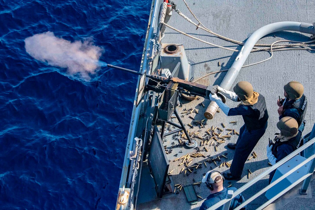 A sailor fires a gun from the deck of a ship as two others watch.