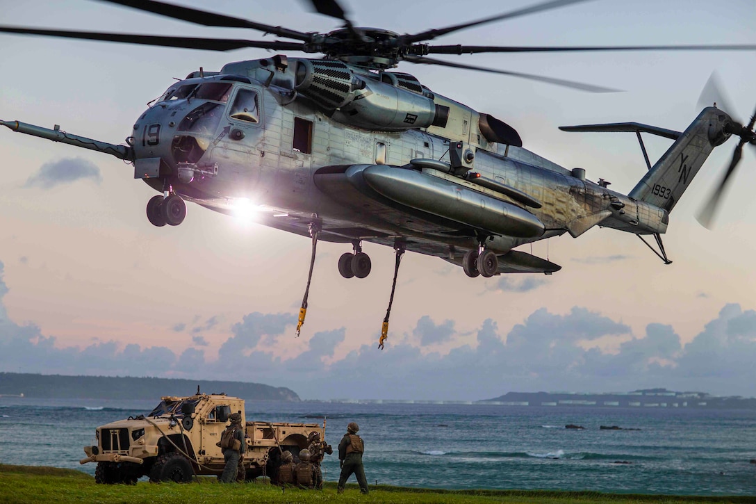 Marines prepare to attach a vehicle to an airborne helicopter.