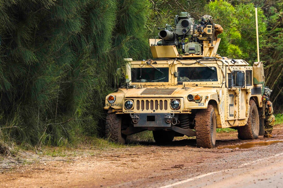 A soldier mans a gun on a military vehicle as another kneels behind the vehicle.