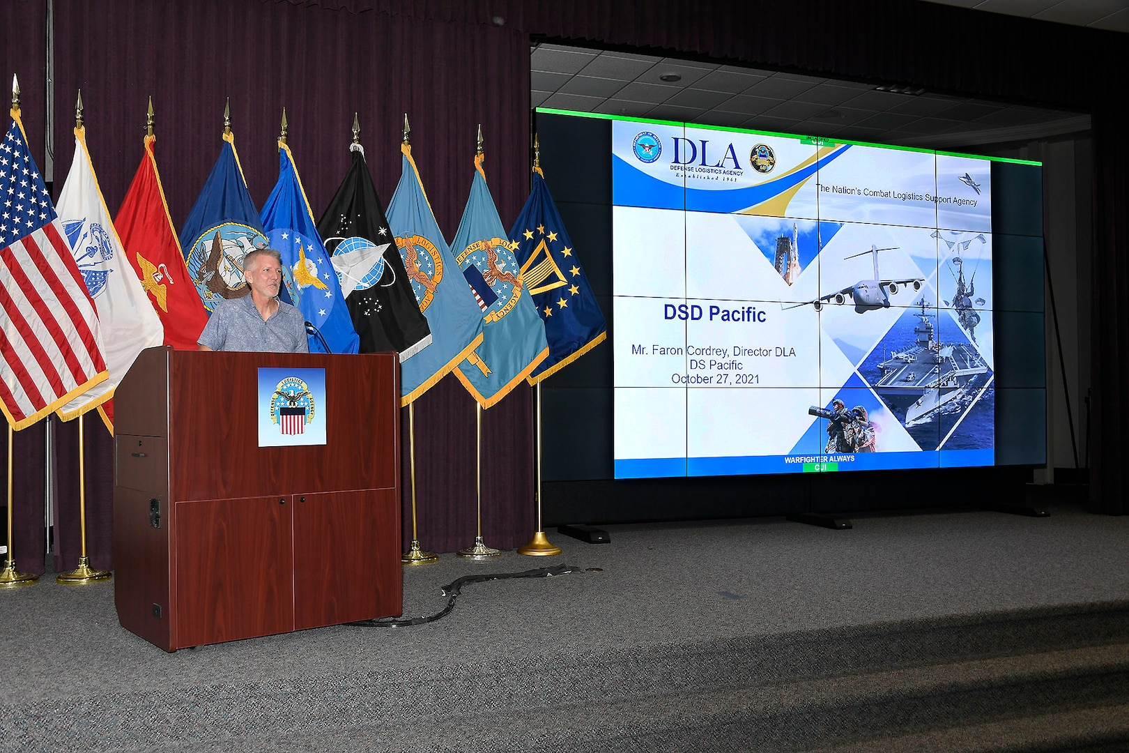 Man standing on stage behind podium with flags in the background.