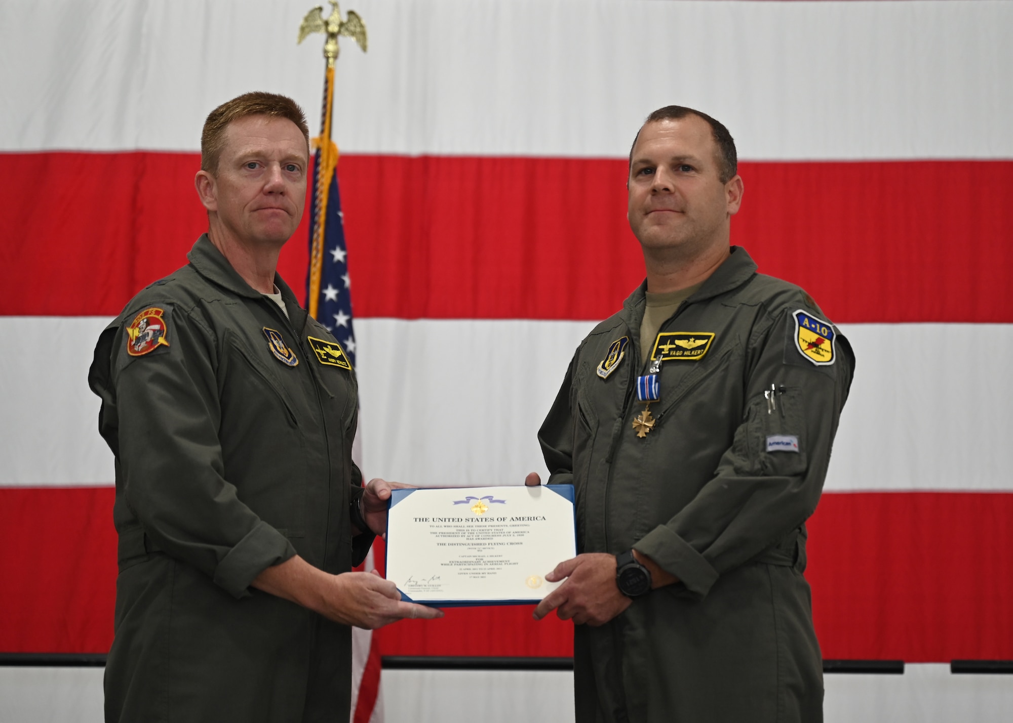 Two men in green coveralls hold a medal citation in front of a large American flag. The citation reads "The United States of America. To all who shall see these presents, greetings. This is to certify that the president of the United States of America, authorized by act of Congress July 5, 1928, has awarded the Distinguished Flying Cross with "v" device to Captain Michael J. Hilkert for extraordinary achievement while participating in aerial flight."