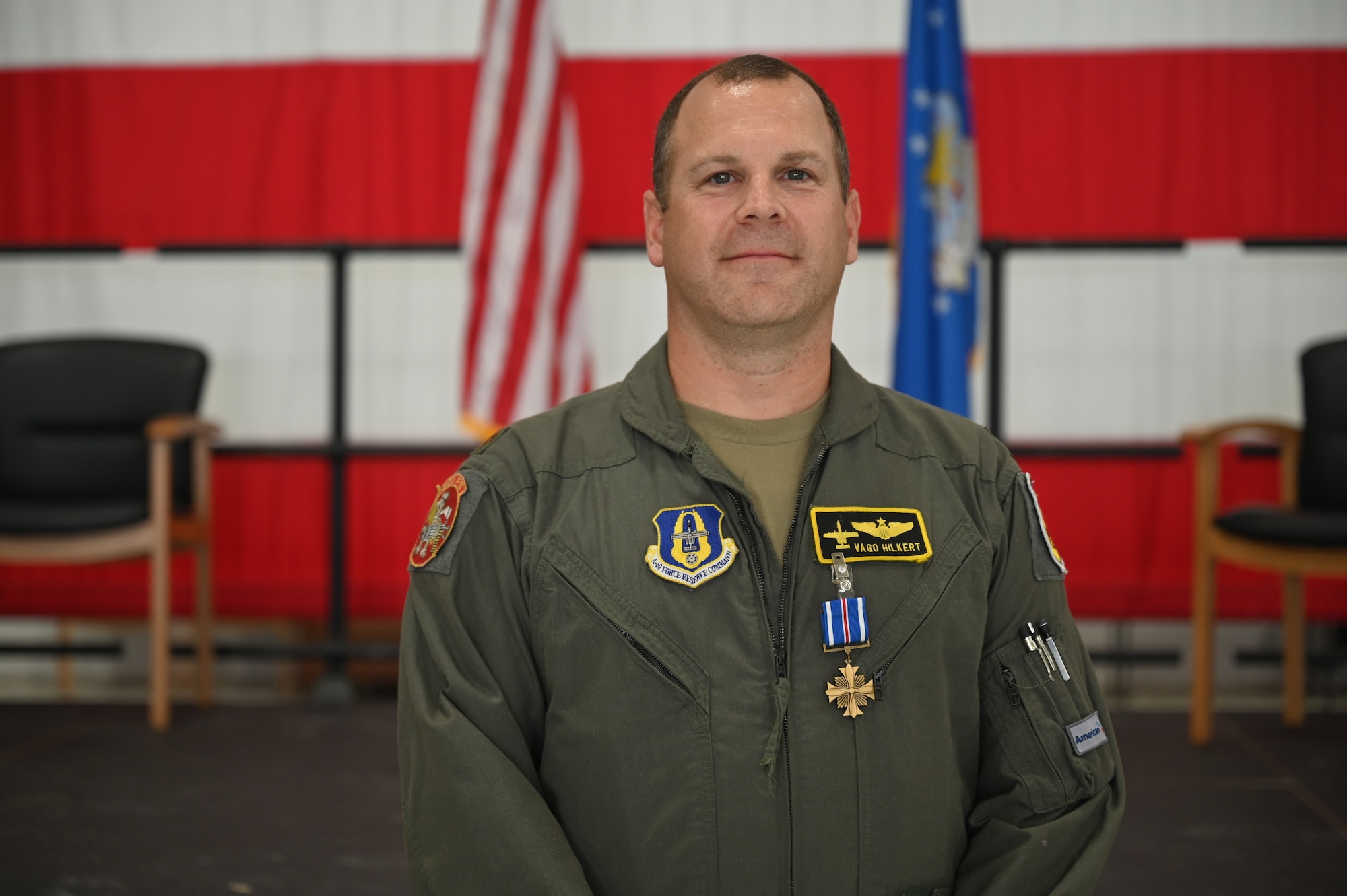 A man stands in front of a large American flag wearing green coveralls. The patch on his left breast reads "Vago Hilkert". A medal is clipped to his name patch.