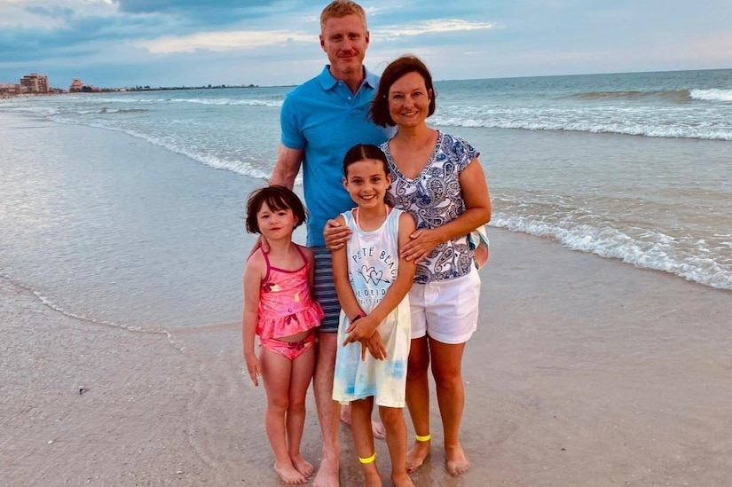 A man and woman stand on a beach with two children; the ocean is in the background.