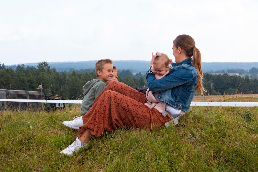 A woman sits on the grass holding a baby; two small children sit to her side.