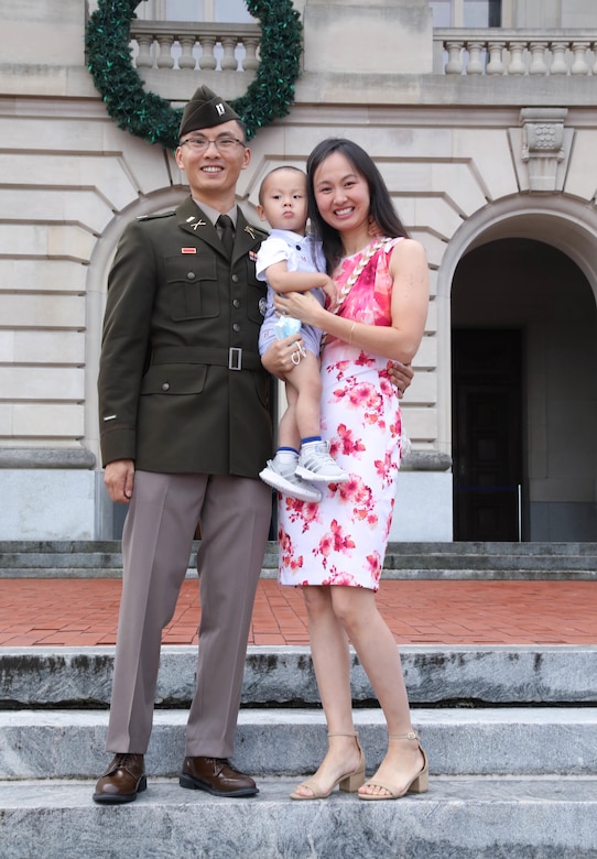 A man wearing a military uniform stands next to a woman holding a child. A building is in the background.