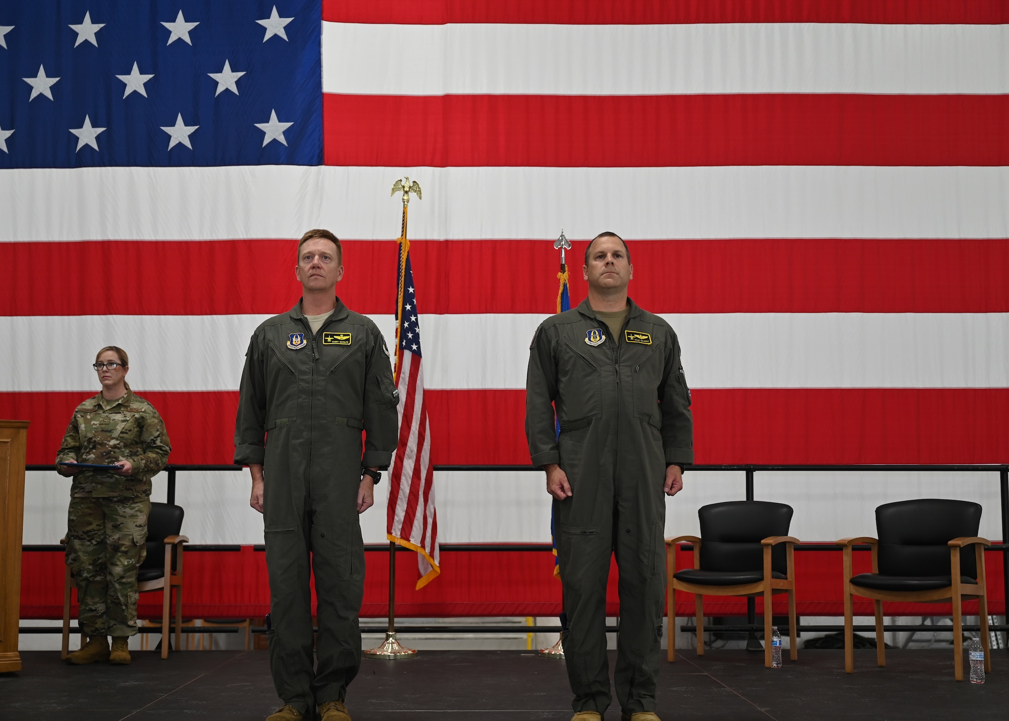 Two men in green coveralls stand at attention in front of a large American flag.