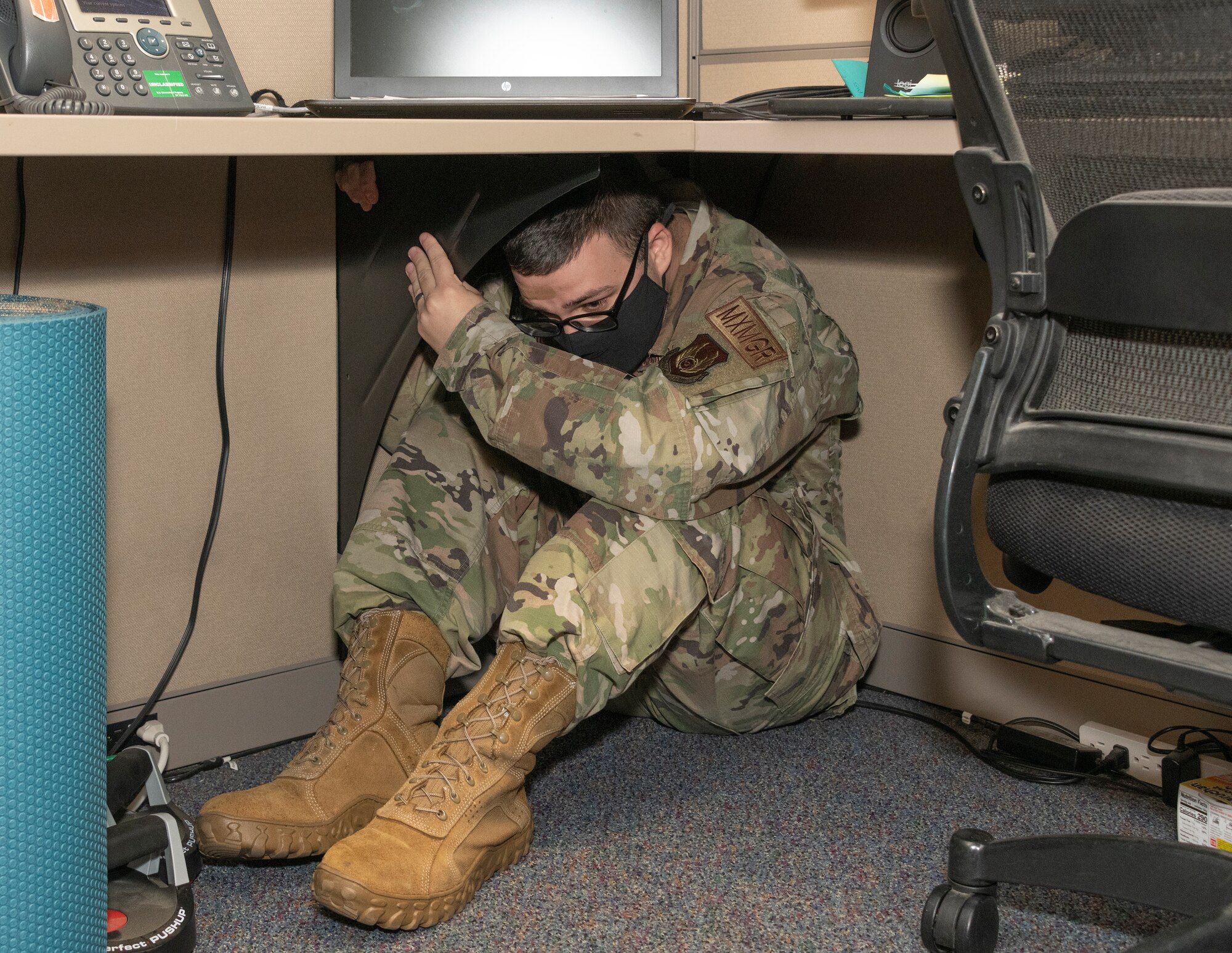 Senior Airman Tyler Rogers, 60th Maintenance Group plans, scheduling and documents, practices drop, cover and hold on under his desk Oct. 21, 2021, at Travis Air Force Base, California. Several units at Travis AFB worked together to practice and improve catastrophic incident response skills during the Great Shake Out earthquake exercise. (U.S. Air Force photo by Heide Couch)