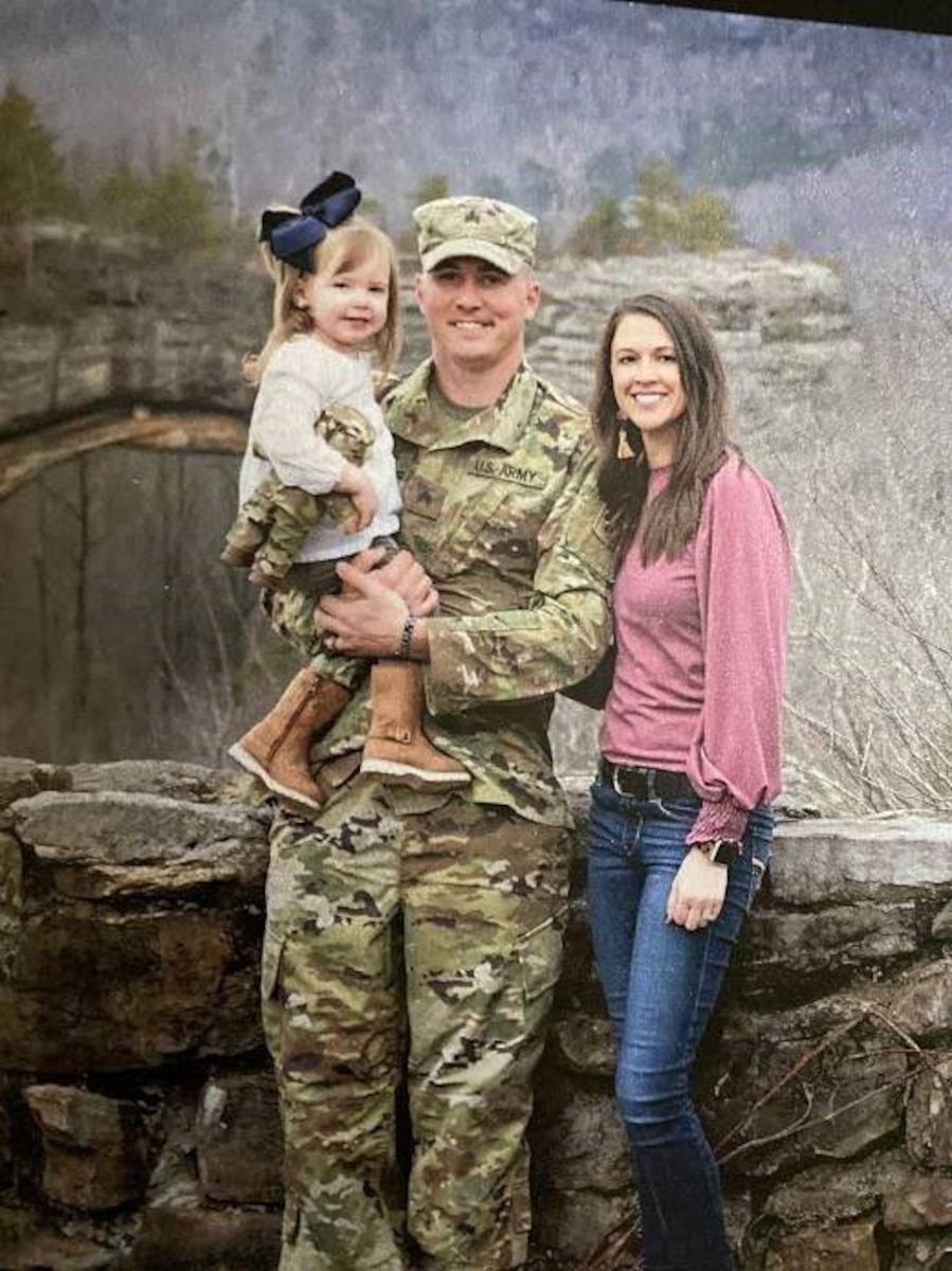 A woman stands next to a man wearing a military uniform, who's holding a child. Mountains and trees are in the background.